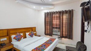 A hotel room with a wooden headboard bed, white bedding, colorful pillows, a desk with a chair, a window with patterned curtains, and a wall-mounted TV. - Via De Gold I (New Delhi)