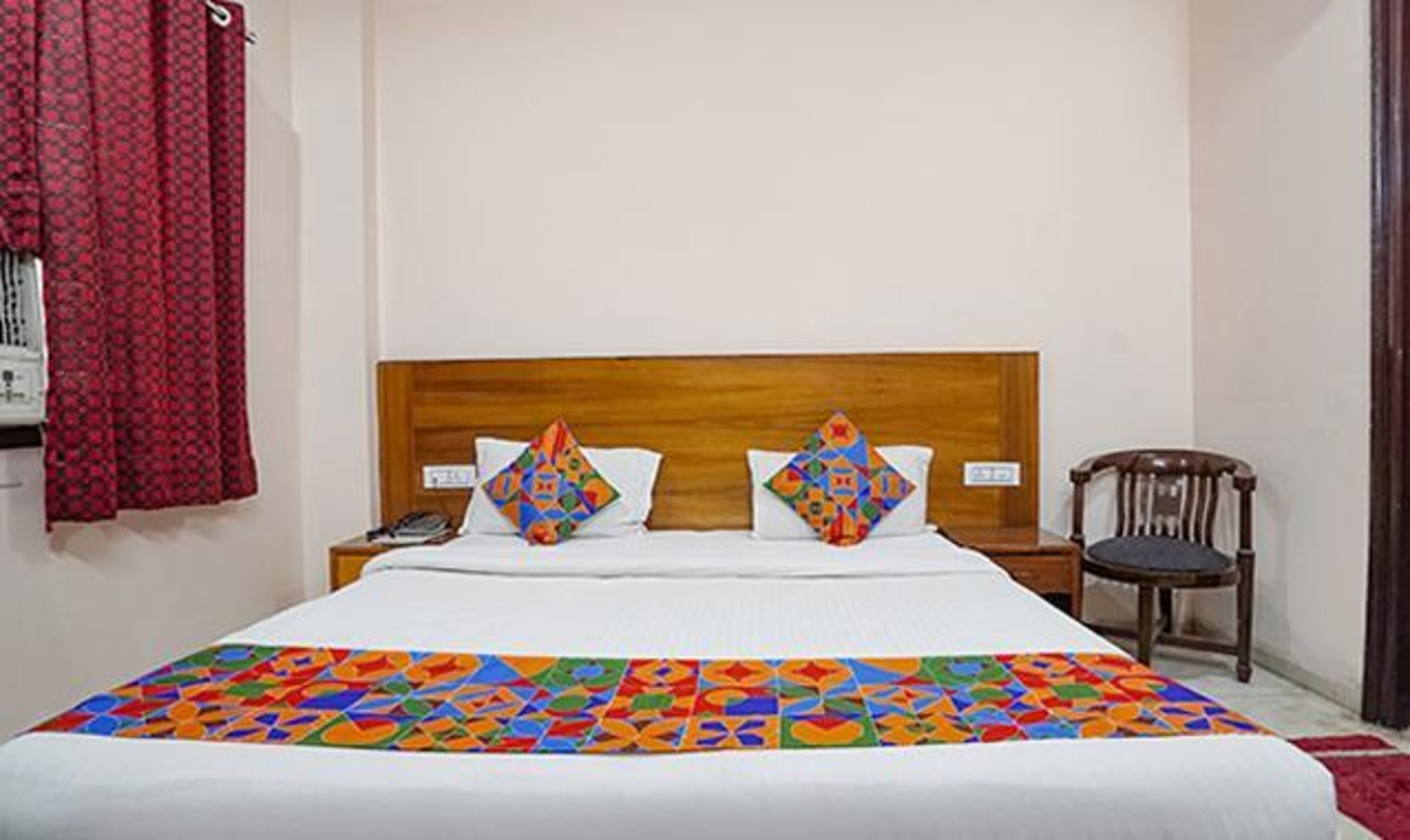 A hotel room with a wooden headboard bed, two colorful pillows, a red curtain, a wooden chair, and a patterned bedspread.