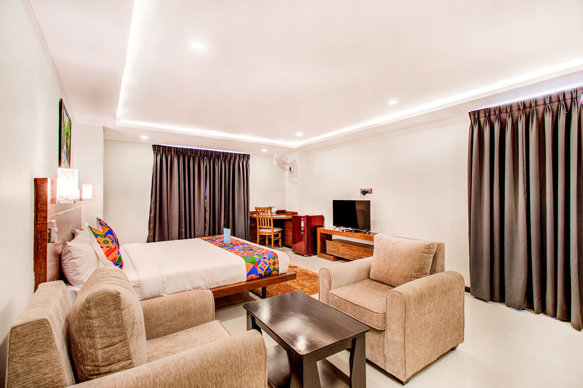 A modern hotel room with a large bed, two armchairs, a small table, a desk with a TV, and a wardrobe.