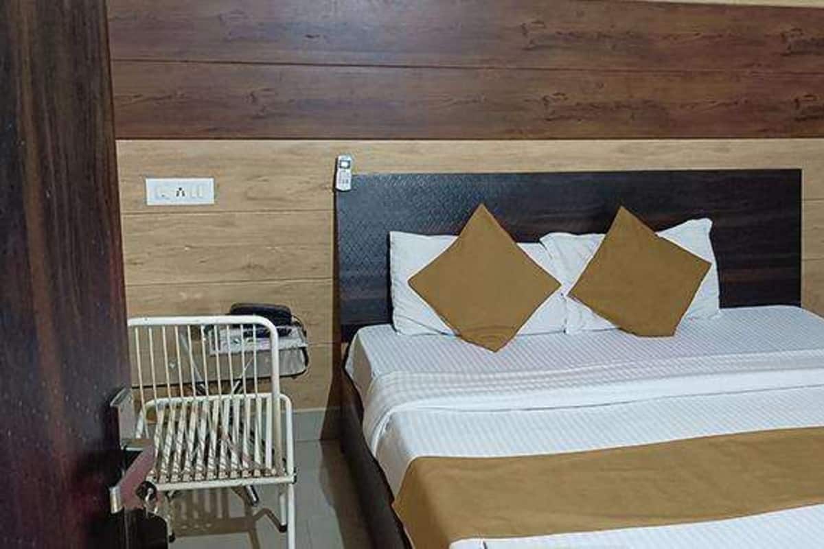 A hotel room with a wooden headboard, a bed with white bedding and two brown pillows, a bedside table with a phone, and a metal chair.