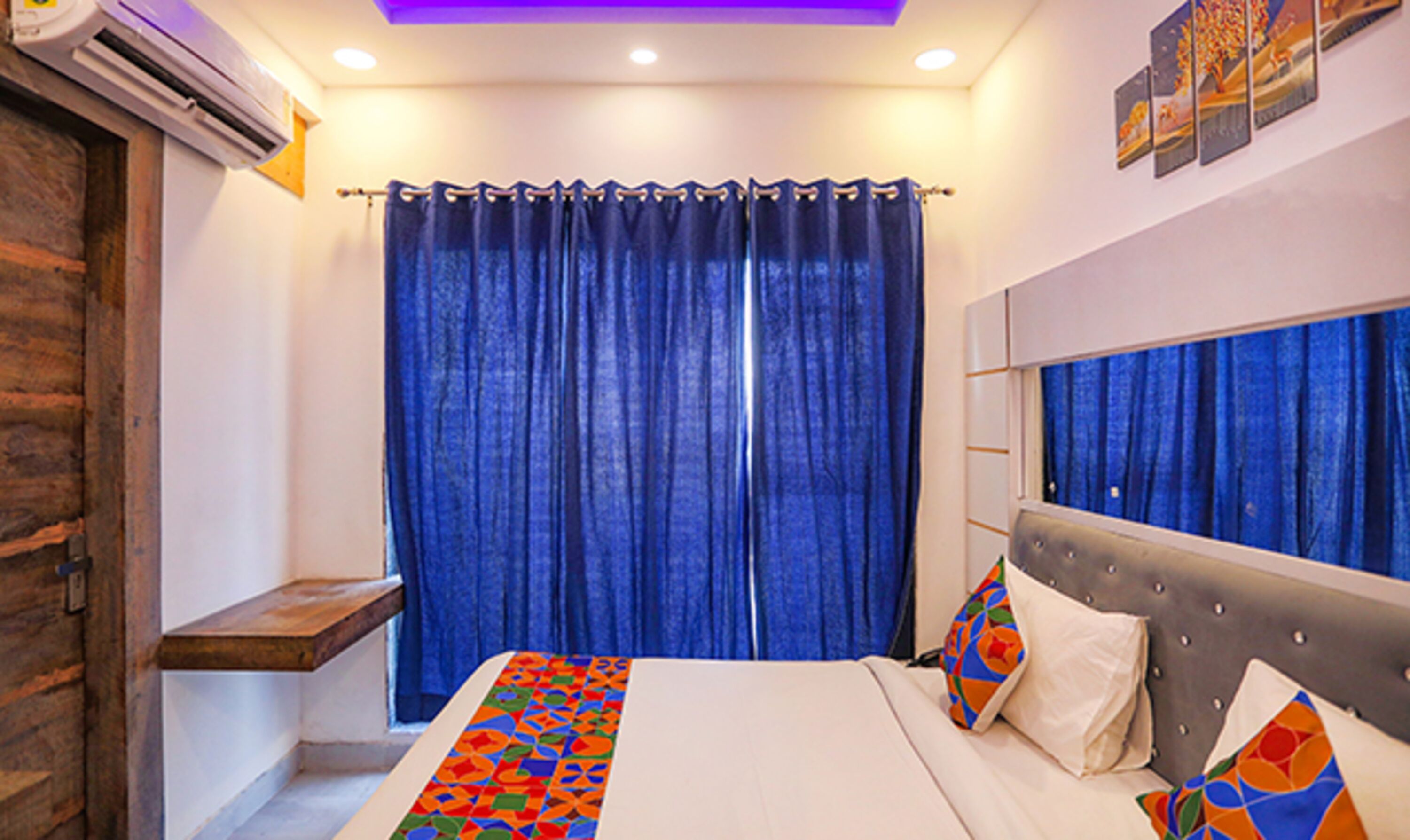 A hotel room with a bed, a bedside table, a window with blue curtains, and a wall-mounted mirror.