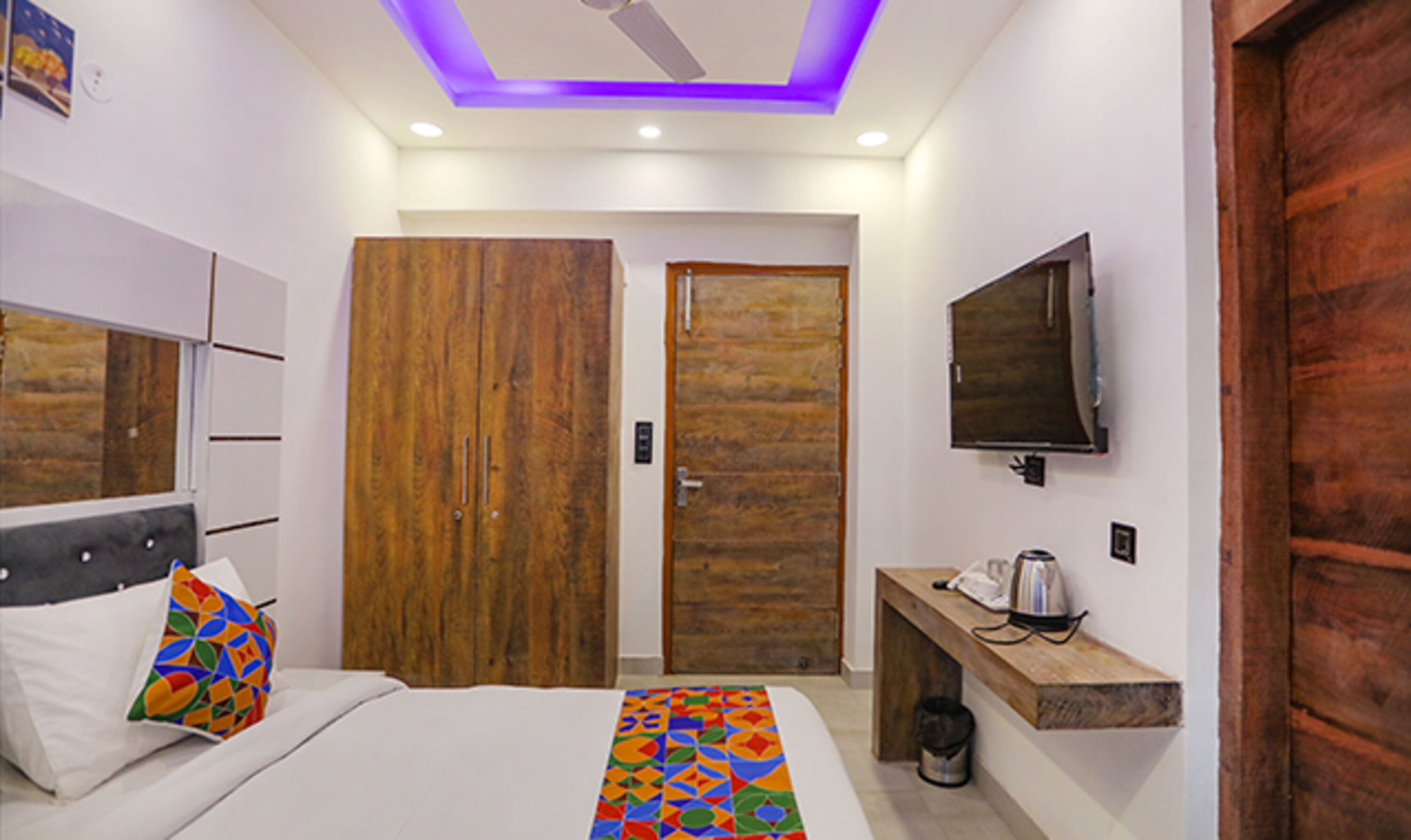 A bedroom with a bed, a wooden wardrobe, a bench with a kettle, and a TV mounted on the wall.