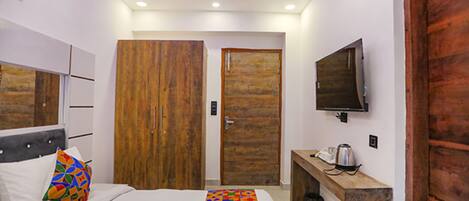 A bedroom with a bed, a wooden wardrobe, a bench with a kettle, and a TV mounted on the wall.