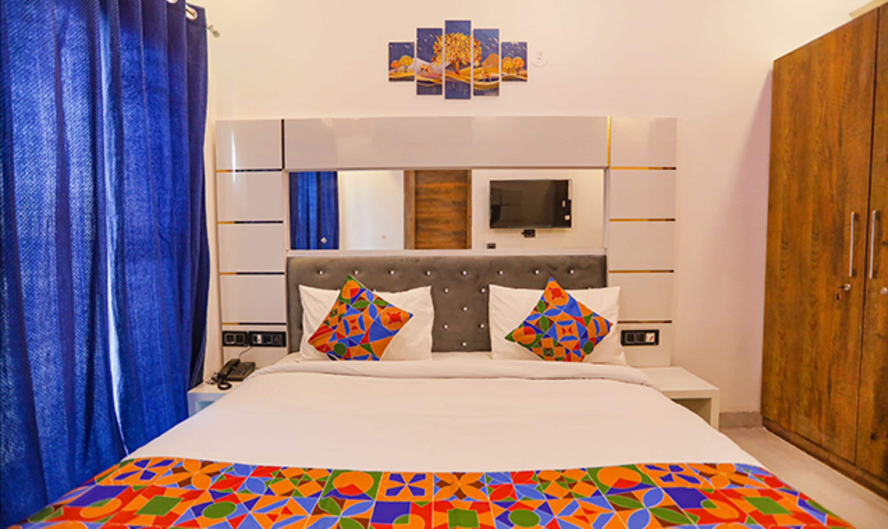 A hotel room with a bed, a TV, a wooden wardrobe, and a colorful geometric-patterned bedspread.