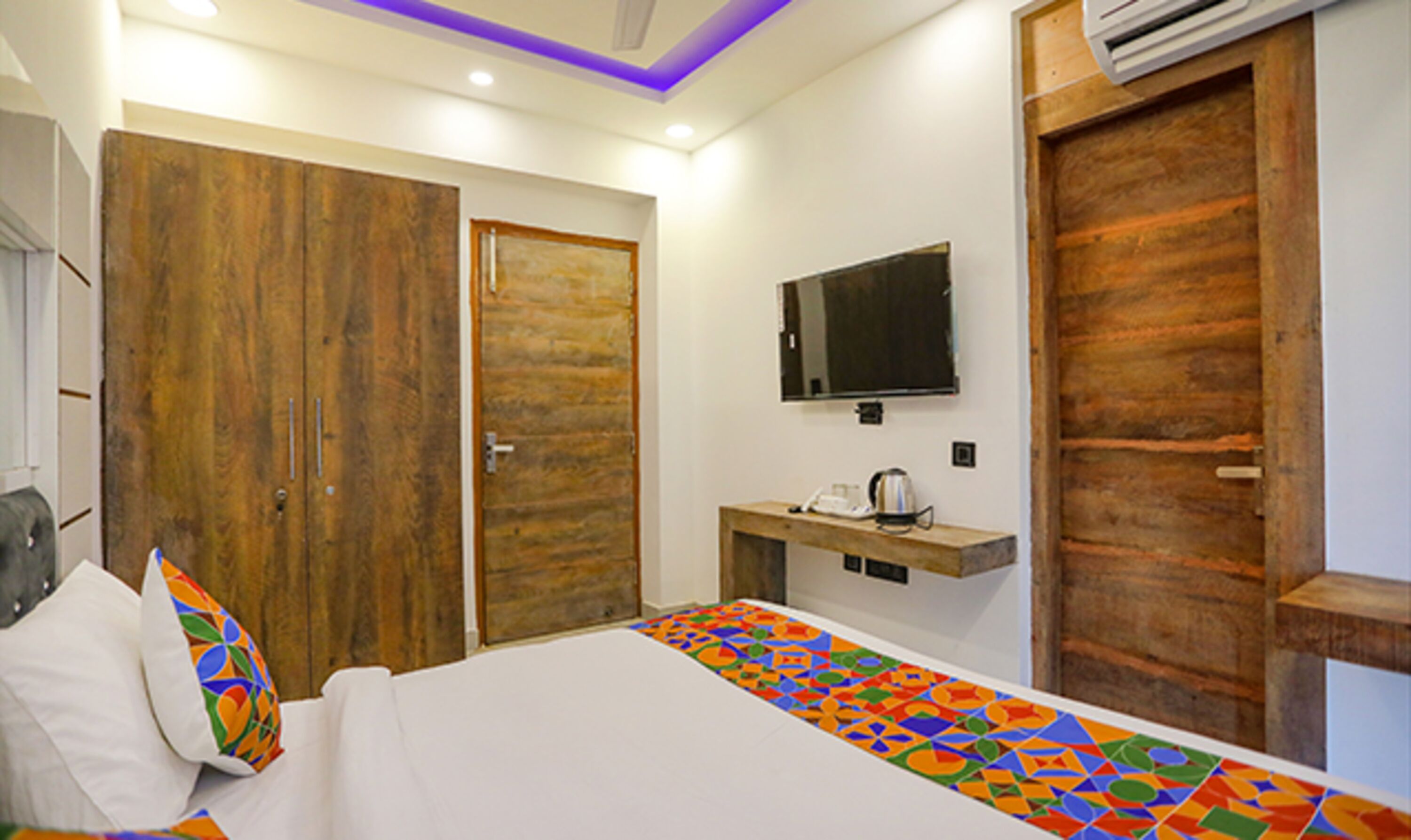 A bedroom with a bed, a TV mounted on the wall, a wooden wardrobe, and a small table with a kettle.
