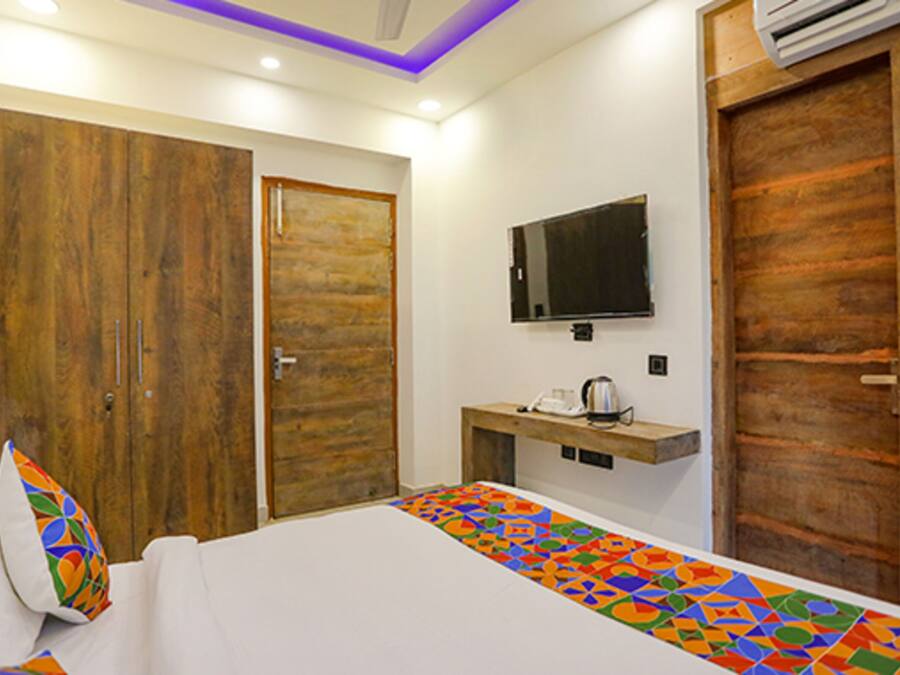 Quarto com cama, televisão na parede, guarda-roupa de madeira e uma pequena mesa com chaleira.