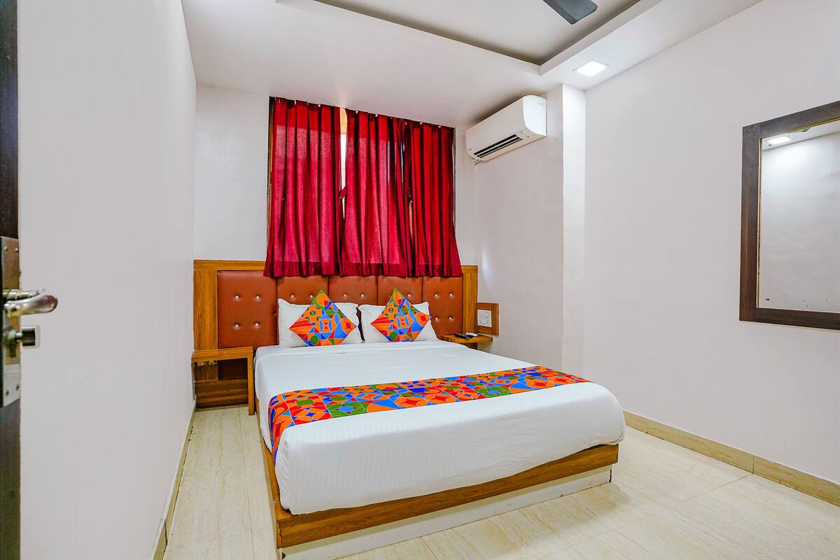 A hotel room with a bed, red curtains, a wooden headboard, a nightstand, and a wall-mounted air conditioning unit.