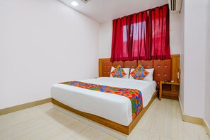A neatly made bed with a colorful bedspread and pillows, a wooden headboard, and a red curtain. - FabExpress Diamond (Mumbai)