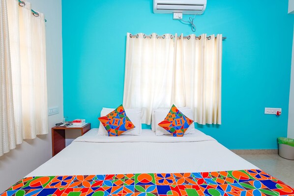 Habitación de hotel con una pared turquesa, una cama con ropa de cama blanca, dos almohadas coloridas, una cortina blanca y una alfombra de colores.
