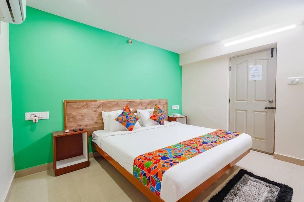 A hotel room with a bed, a nightstand, a wall socket, a door, and a light switch.