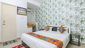 Quarto com cama, televisão fixada na parede, papel de parede estampado e uma pequena mesa com cadeira.