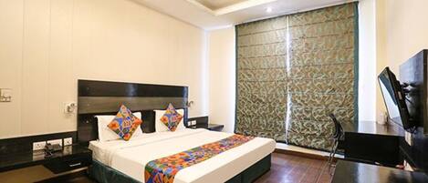 A hotel room with a bed, a desk, a television, and a window with patterned curtains.
