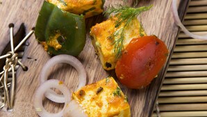Uma tábua de madeira com cubos de queijo paneer, tomates, cebolas e um pimentão verde.