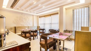 Espaço de refeições moderno em hotel, com mesa central, várias cadeiras e um painel decorativo na parede.