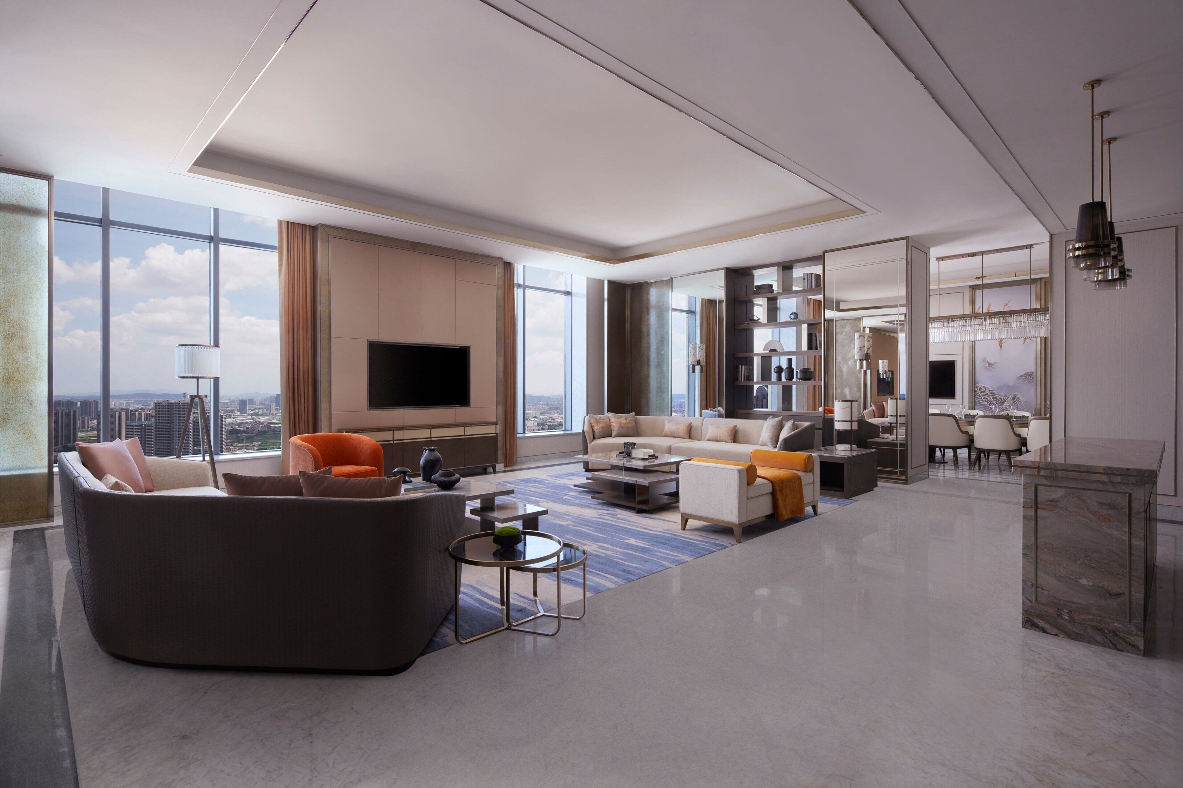A spacious living room with a large sofa, coffee table, and a flat-screen TV. There is a dining area with chairs and a chandelier. The room has large windows offering a city view.