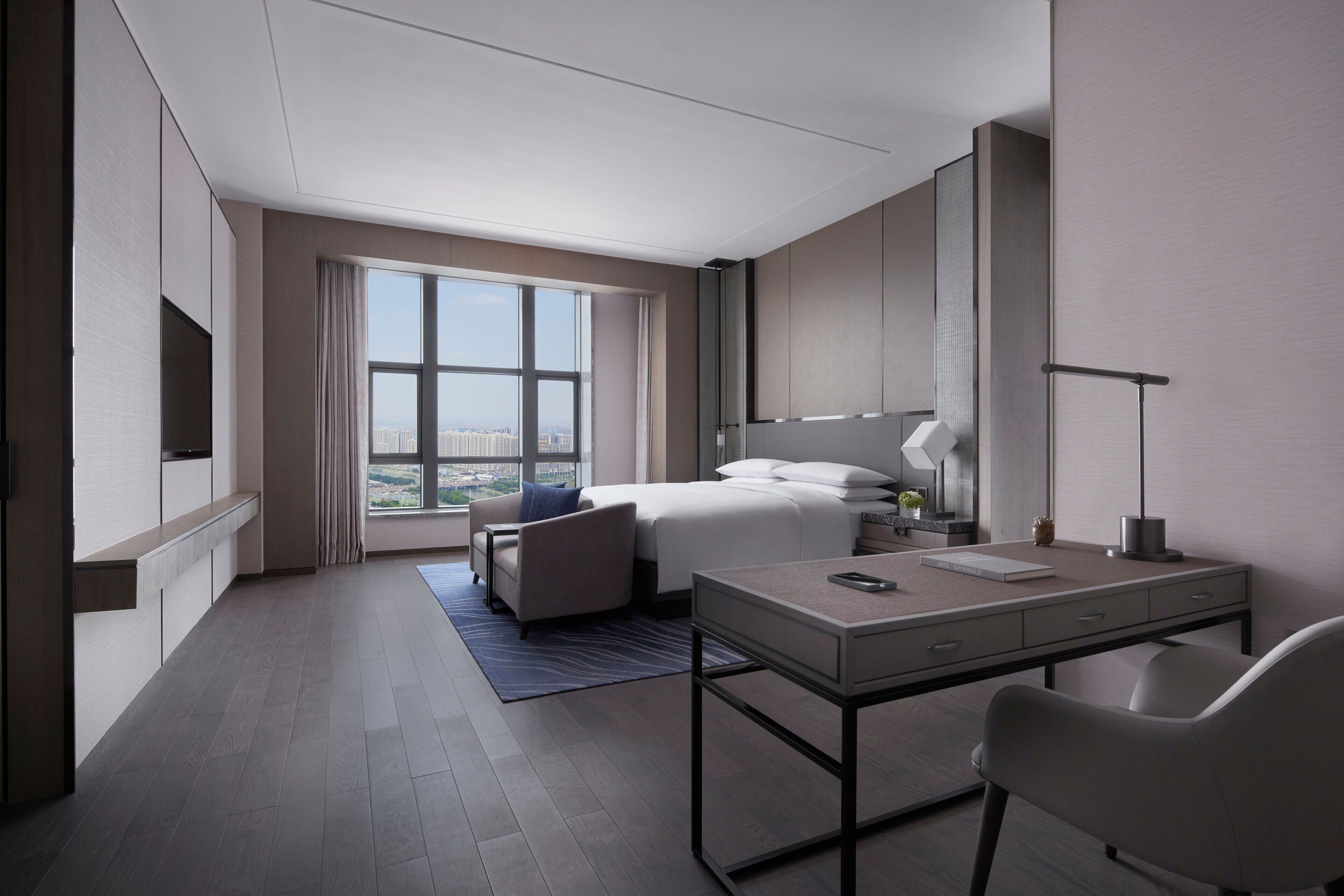 A modern hotel room with a large bed, a desk, a chair, a TV, and a view of the city.