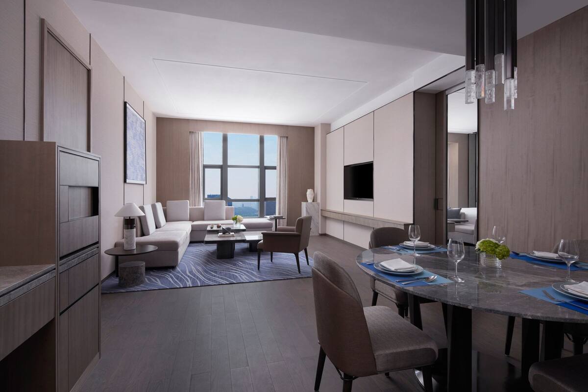 Quarto de hotel moderno com área de jantar, um sofá, uma televisão de tela plana e uma janela grande com vista.