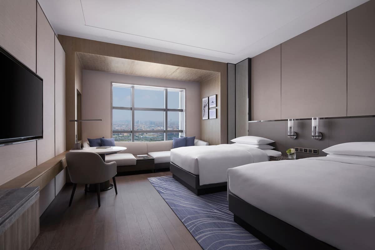 Quarto de hotel moderno com uma janela grande, duas camas, uma TV de tela plana, área de estar e vista para a cidade.