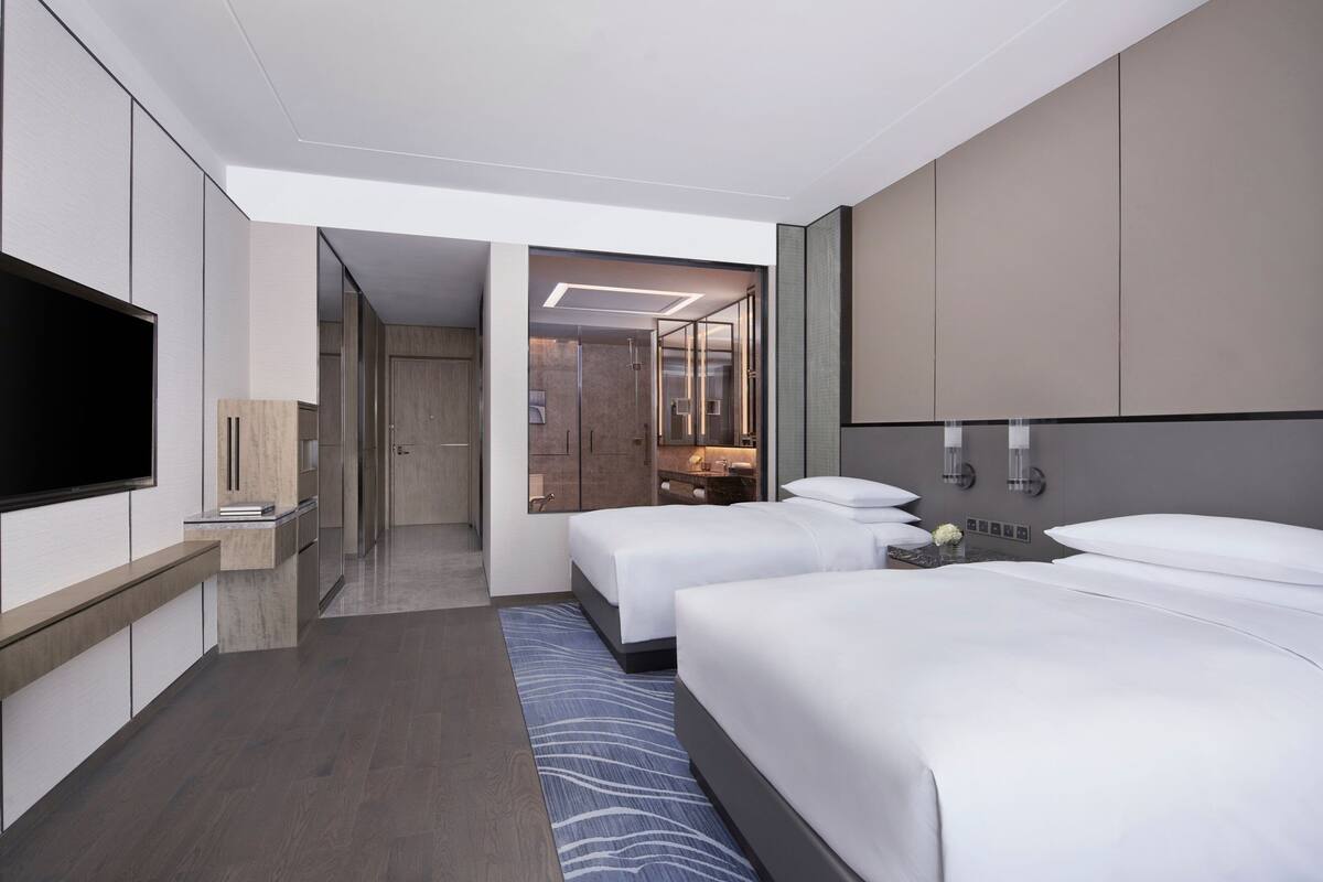 Quarto de hotel moderno com duas camas, televisão de tela plana, banheiro com chuveiro de vidro e closet espaçoso.