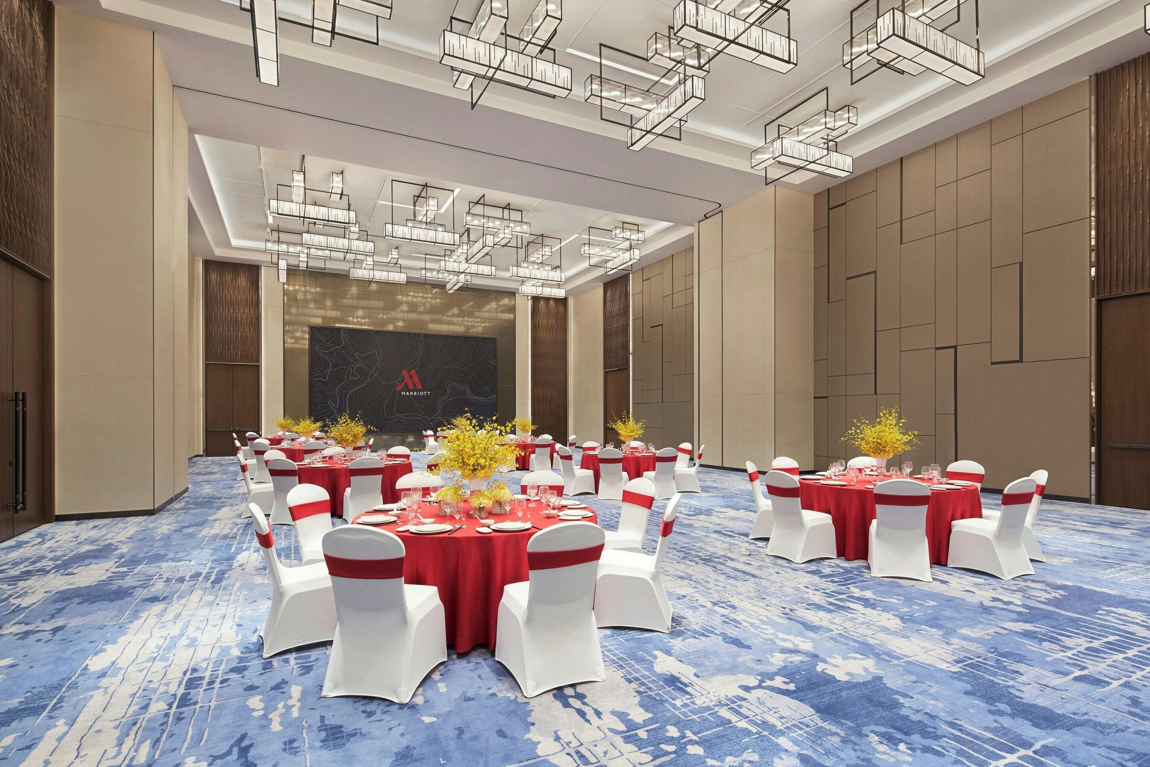 A banquet hall with round tables set for dining, white chairs with red sashes, and a blue patterned carpet.