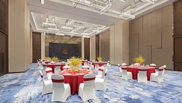 A banquet hall with round tables set for dining, white chairs with red sashes, and a blue patterned carpet.