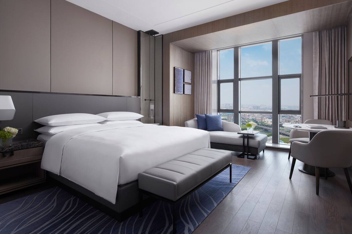 Quarto de hotel moderno com uma cama grande, um sofá, uma mesinha e vista da cidade através de grandes janelas.