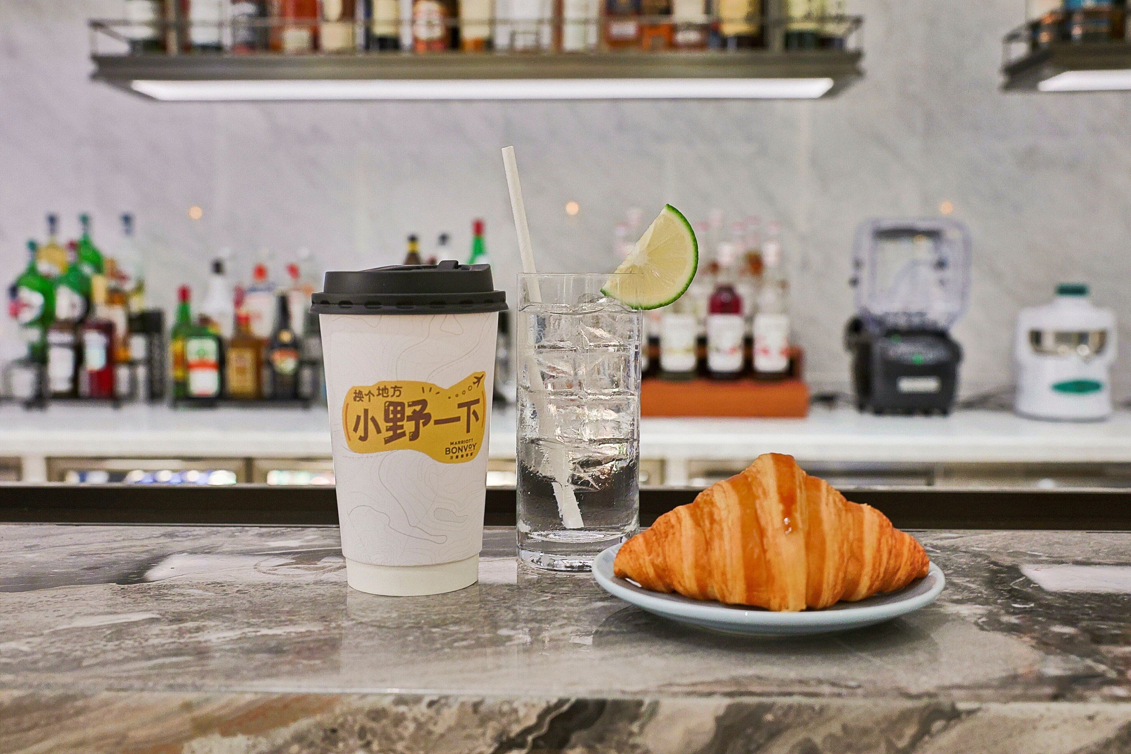 Ein Trinkbecher, ein Glas mit Limettenscheibe und ein Croissant auf einer Marmorplatte.