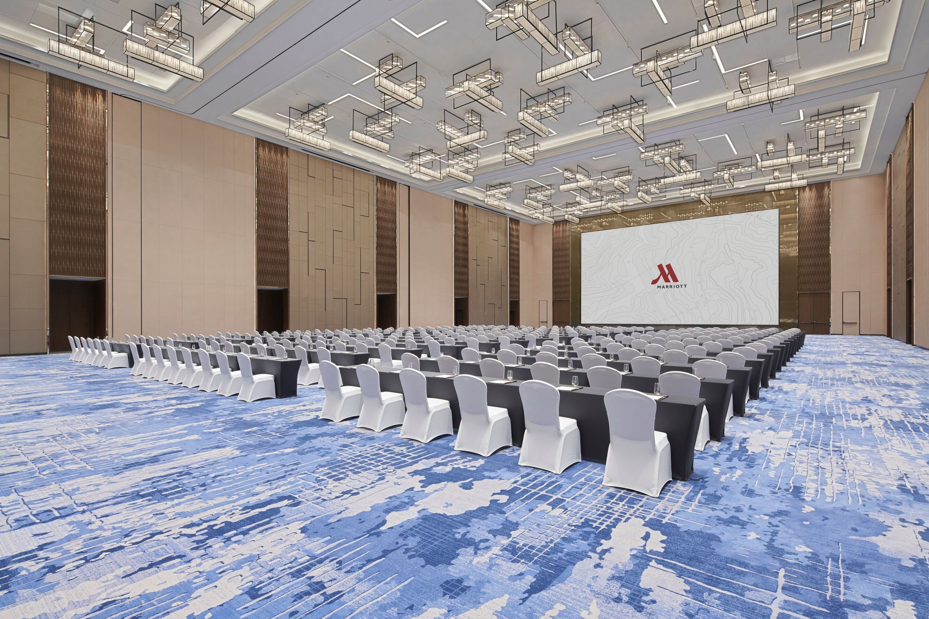 A large conference room with rows of chairs, a stage with a screen, and a modern ceiling design.