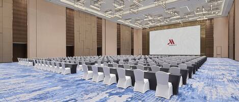 A large conference room with rows of chairs, a stage with a screen, and a modern ceiling design.