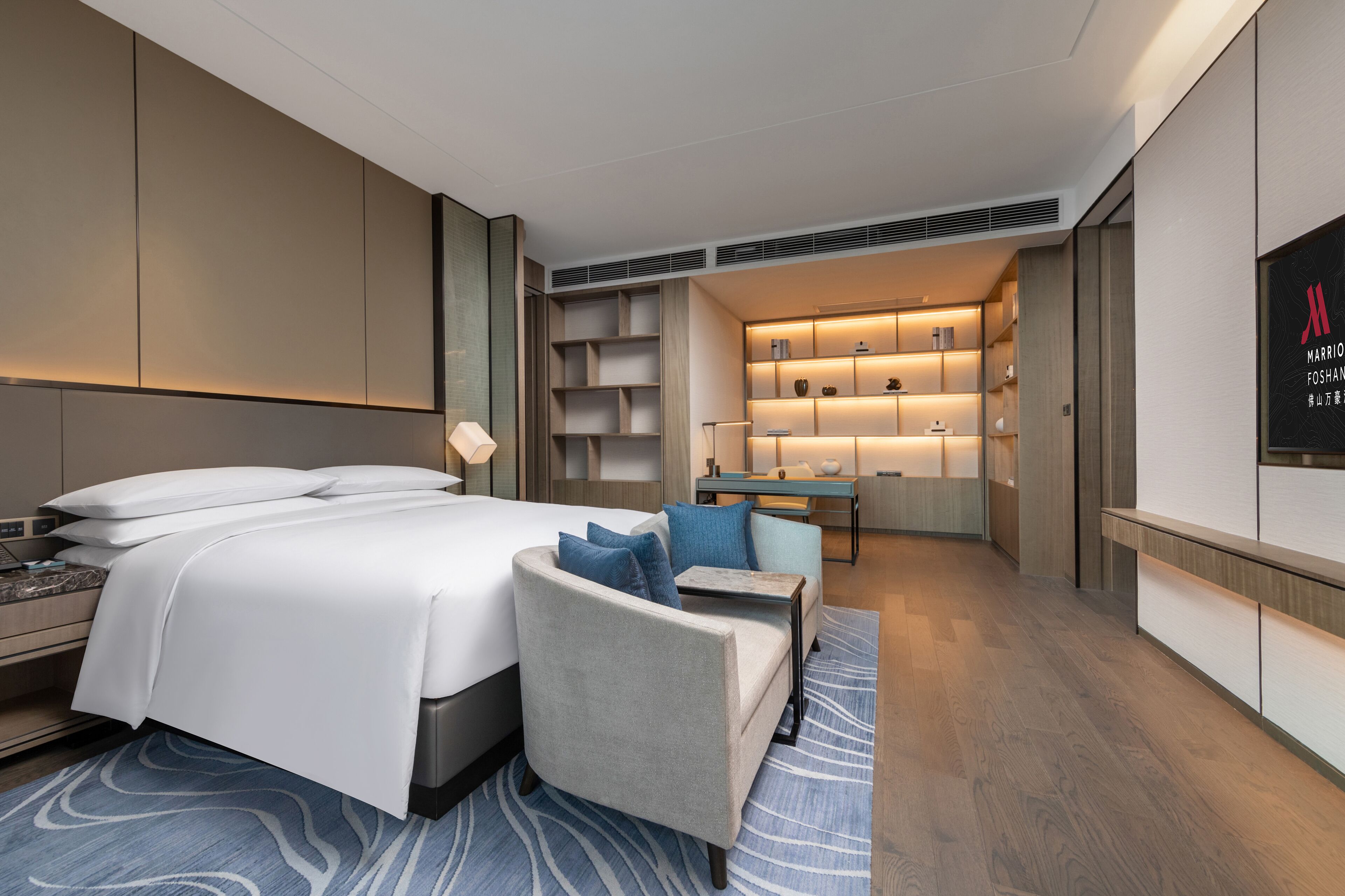 A modern hotel room with a large bed, a seating area with a table, and built-in shelving with lighting.
