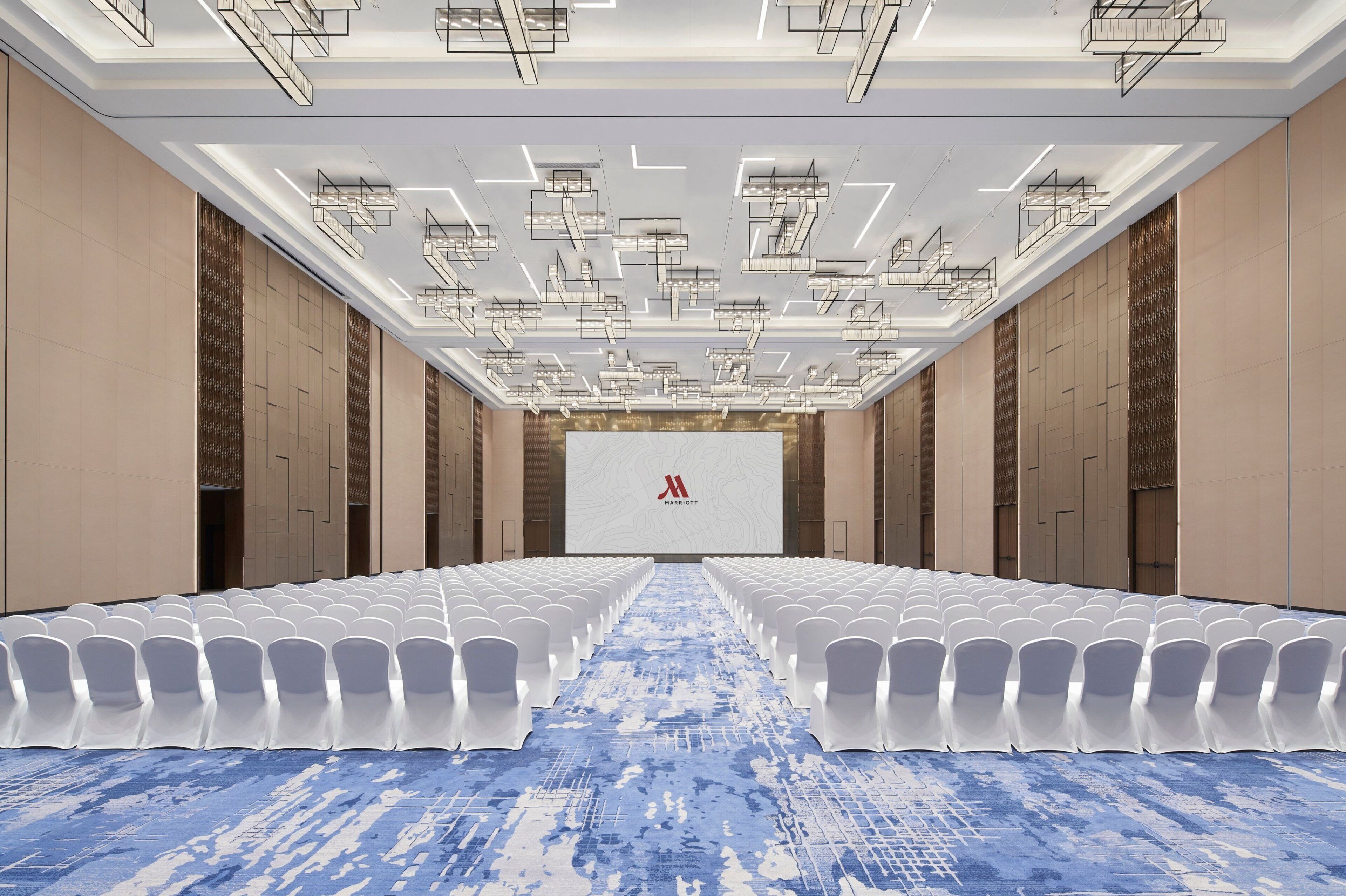 A conference room with rows of white chairs, a blue carpet, and a large screen with a logo.
