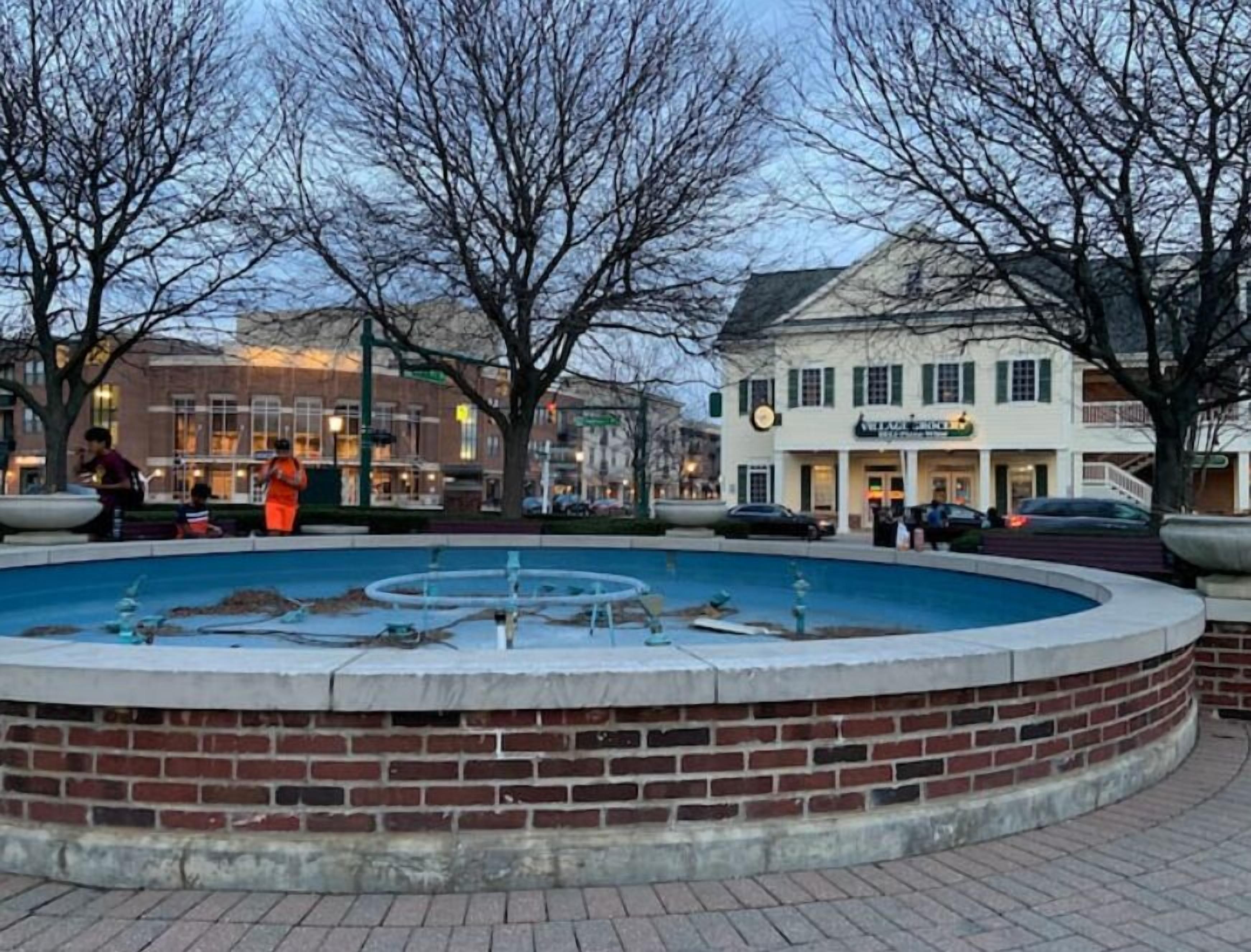 A public fountain with a circular brick base, surrounded by trees and a building with a sign that reads "VILLAGE PLACE."