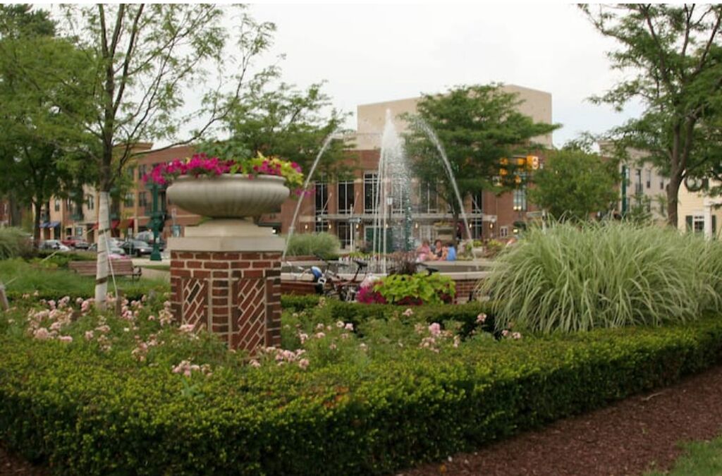 A garden with a fountain, a brick planter with flowers, and a building in the background.