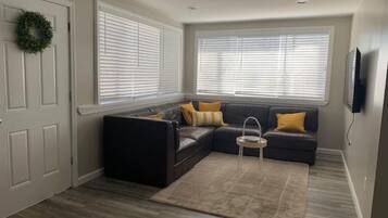 Ein Wohnzimmer mit einem Ecksofa, einem kleinen Beistelltisch und einem großen Fenster mit Jalousien.