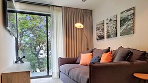 A modern living room with a sofa, a small wooden table, and a view of trees through a large window. - Embrace the Now: 203 Industrial's Sunlit Serenity Awaits (Mexico City)