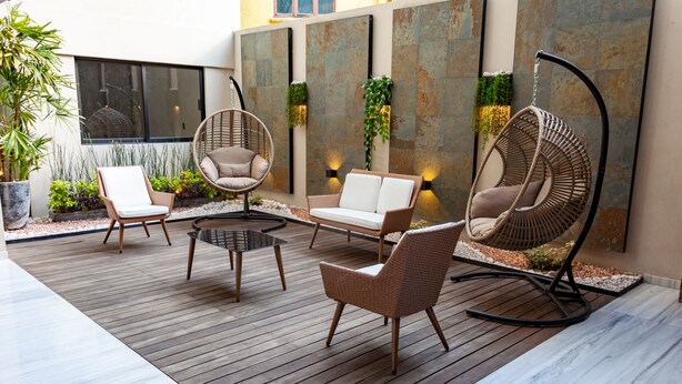 A modern outdoor seating area with wicker chairs and a hanging chair, surrounded by plants and a stone wall.