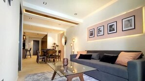 A modern living room with a glass coffee table, grey sofa, and a dining area in the background.