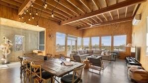A spacious room with a wooden dining table, chairs, a sofa, and a TV. Large windows offer a view of the outdoors. - Luxury Riverfront Retreat | 6 Cabins | Private Pool (Llano)