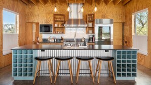 A modern kitchen with a central island, bar stools, wooden cabinetry, and a stainless steel range hood. - Luxury Riverfront Retreat | 6 Cabins | Private Pool (Llano)