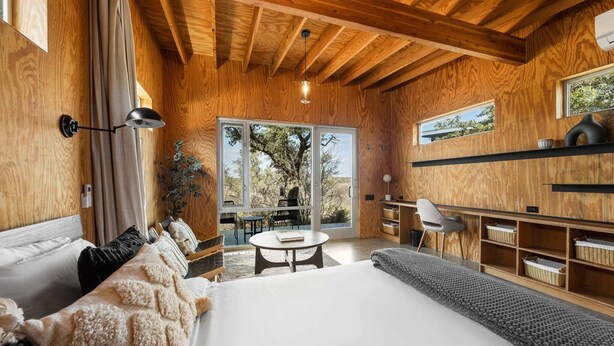 A modern bedroom with a large bed, a small round table, and a view of the outdoors through large windows. - Luxury Riverfront Retreat | 6 Cabins | Private Pool (Llano)