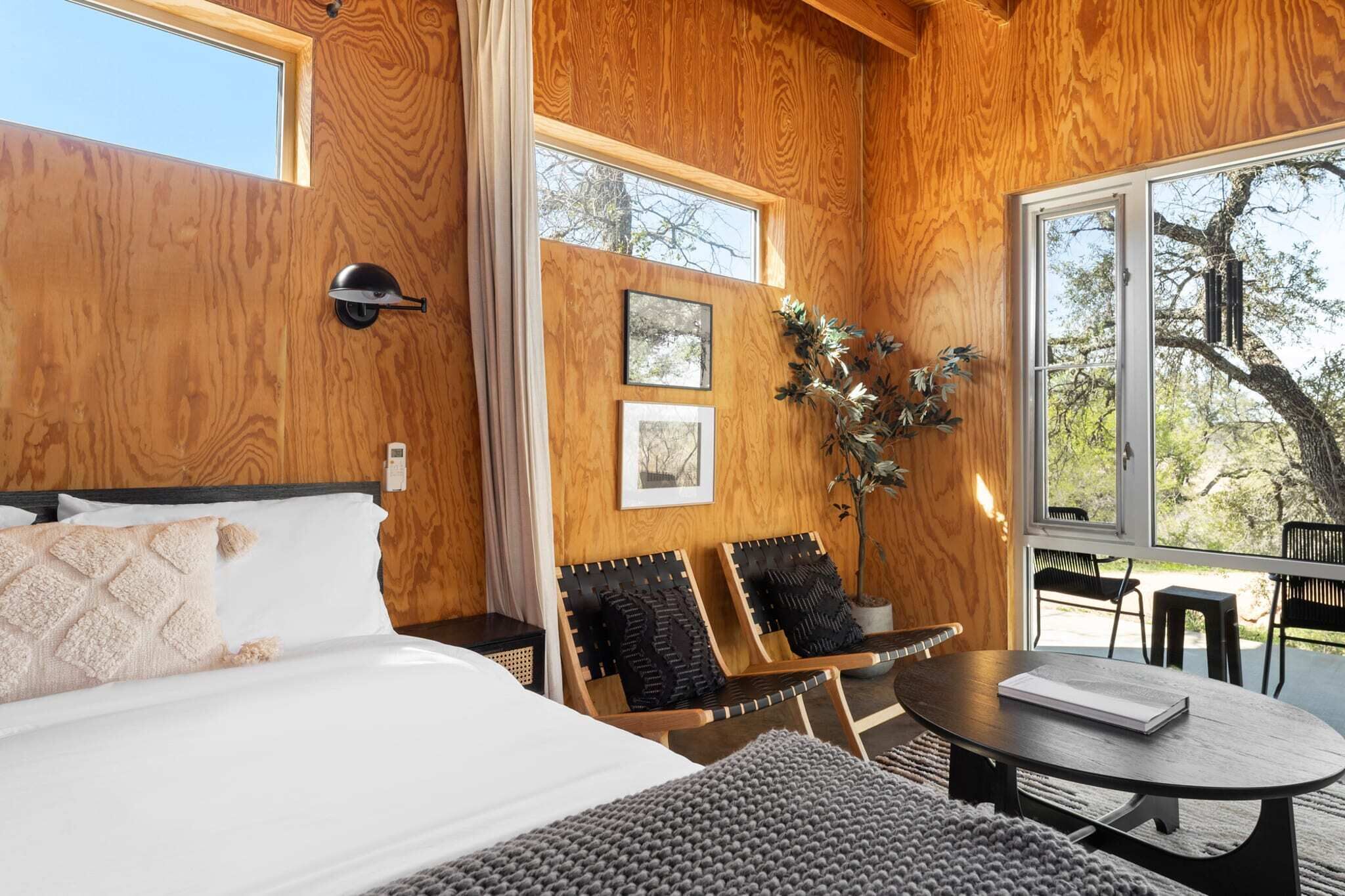 A wooden interior with a bed, two chairs, a table, and a window overlooking a tree.