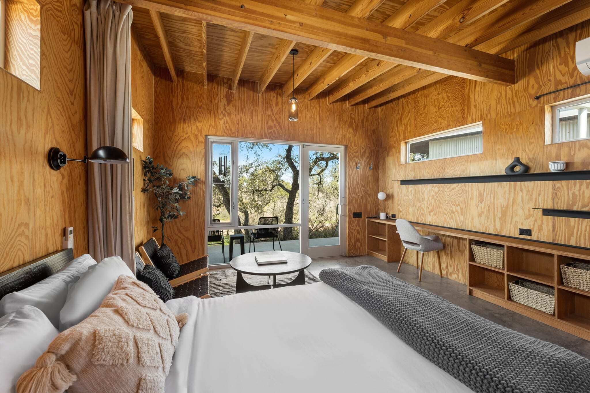 A modern room with a large bed, a desk, and a view of trees through large windows.