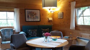 A cozy cabin interior with a round wooden table, four chairs, a sofa, and a ceiling lamp. - Idyllisches Kleines Blockhaus in Großer Gepflegter Gartenlage (Lutterbek)