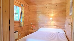 A wooden cabin room with a bed, a window with curtains, a bedside lamp, and a small table with a vase of flowers. - Idyllisches Kleines Blockhaus in Großer Gepflegter Gartenlage (Lutterbek)