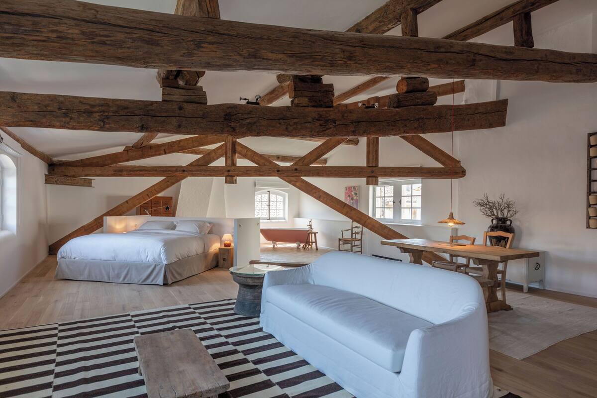 Un espacioso dormitorio tipo loft con vigas de madera a la vista, una cama grande, un sofá y una zona de comedor con vistas a la ciudad.