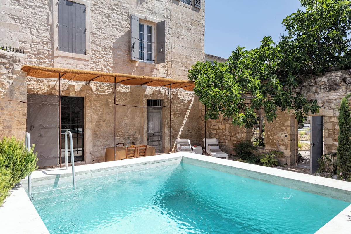 Una piscina con un edificio de piedra al fondo, una zona de estar cubierta y un árbol.