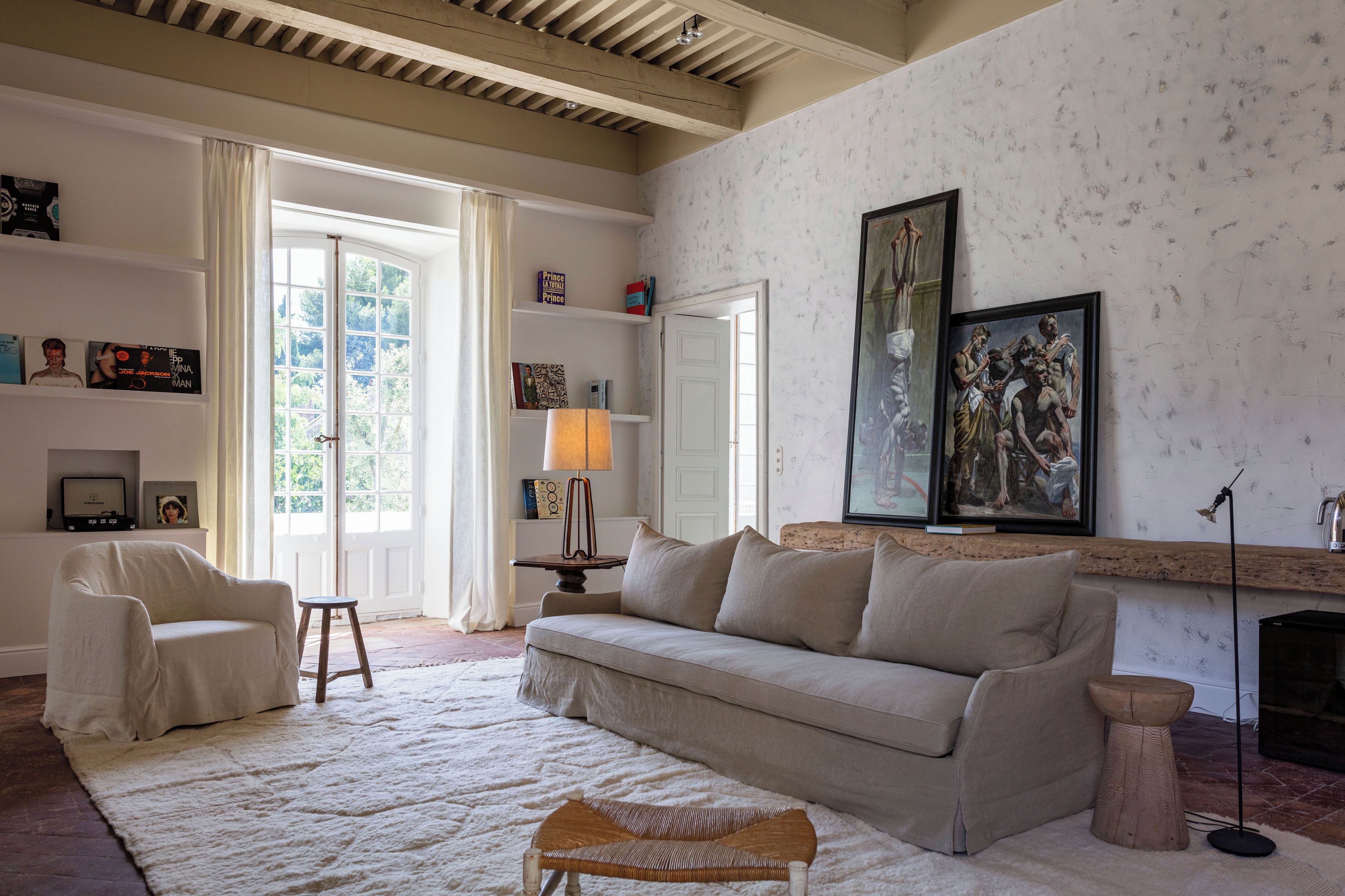 Een woonkamer met een beige bank, een witte fauteuil, een klein houten krukje en een wit vloerkleed. Er hangen ingelijste foto's aan de muur en staat een lamp op een bijzettafeltje.