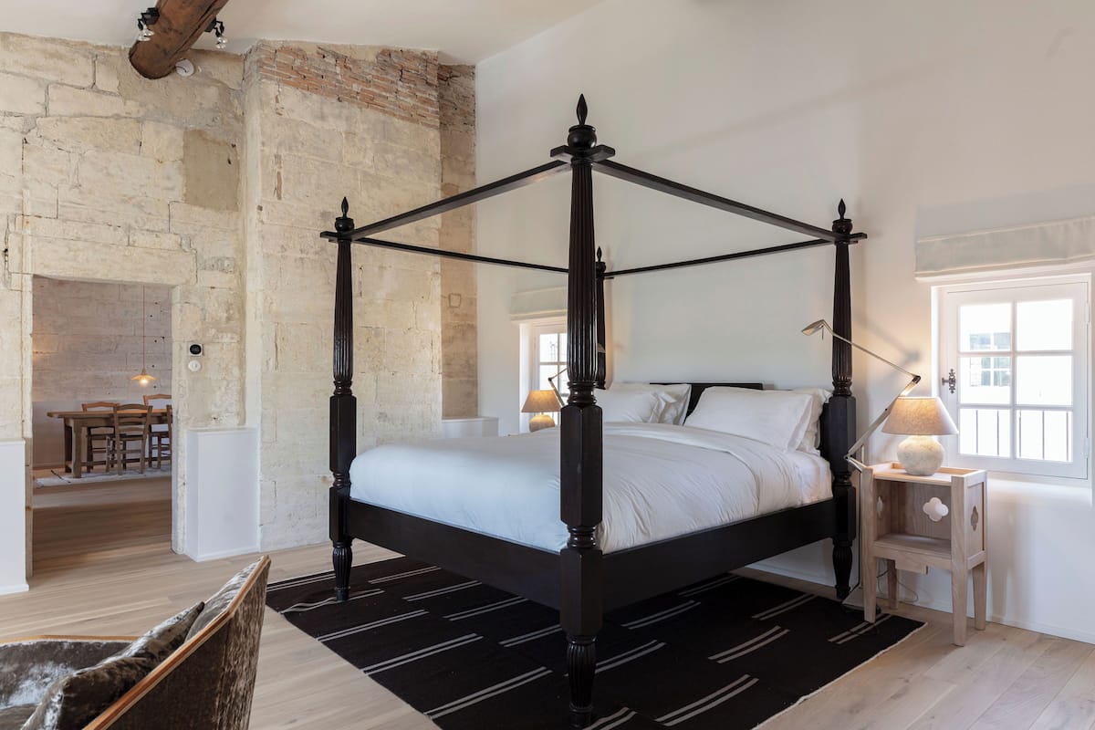 Una cama con dosel en una habitación con paredes de ladrillo visto, piso de madera y una mesita de noche con una lámpara.