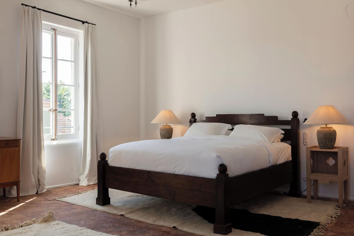 Un dormitorio con una cama de madera, dos lámparas de noche, un ventanal con cortinas y una mesita de noche de madera.