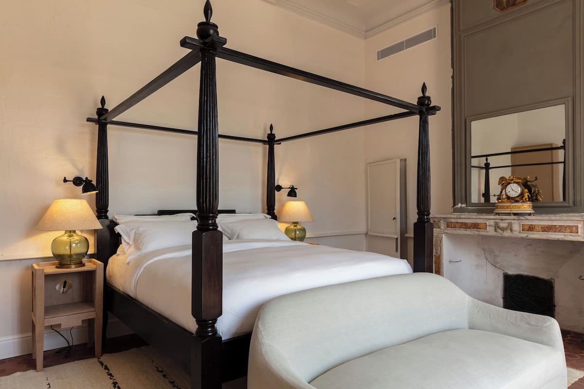 Una cama con dosel, ropa de cama blanca, dos lámparas de noche, una mesita de noche, una silla y una chimenea con repisa.